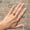 A close-up of a hand with light pink manicured nails wearing a yellow gold ring with a round-cut diamond, set against a blurred cobblestone background.