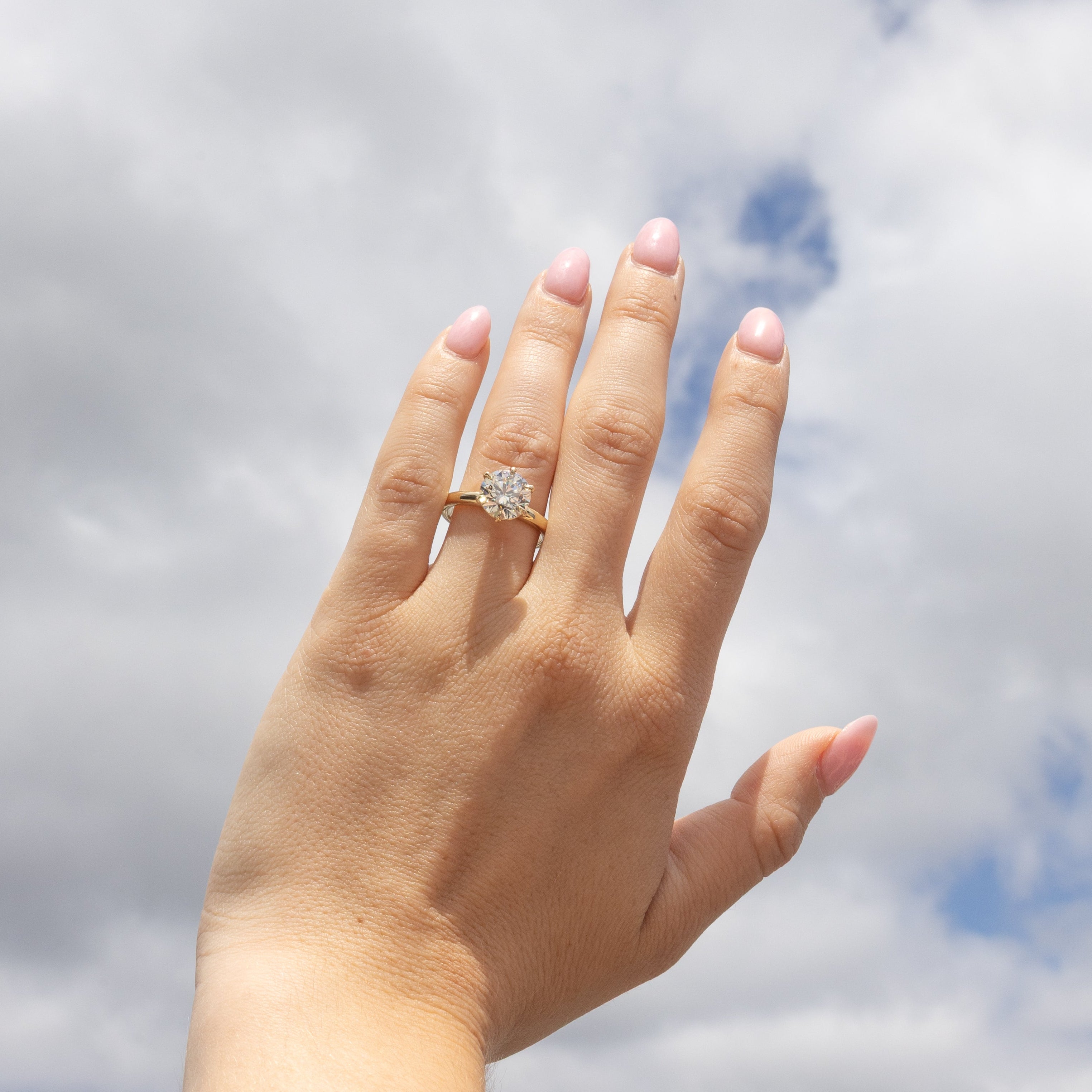 A close-up of a person's hand with pink manicured nails, wearing a gold ring with a round diamond center stone, set against a cloudy blue sky background.