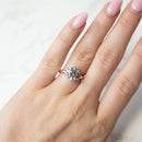 A close-up of a hand with light pink manicured nails wearing a yellow gold ring with a clear round-cut diamond, set against a light marble background.