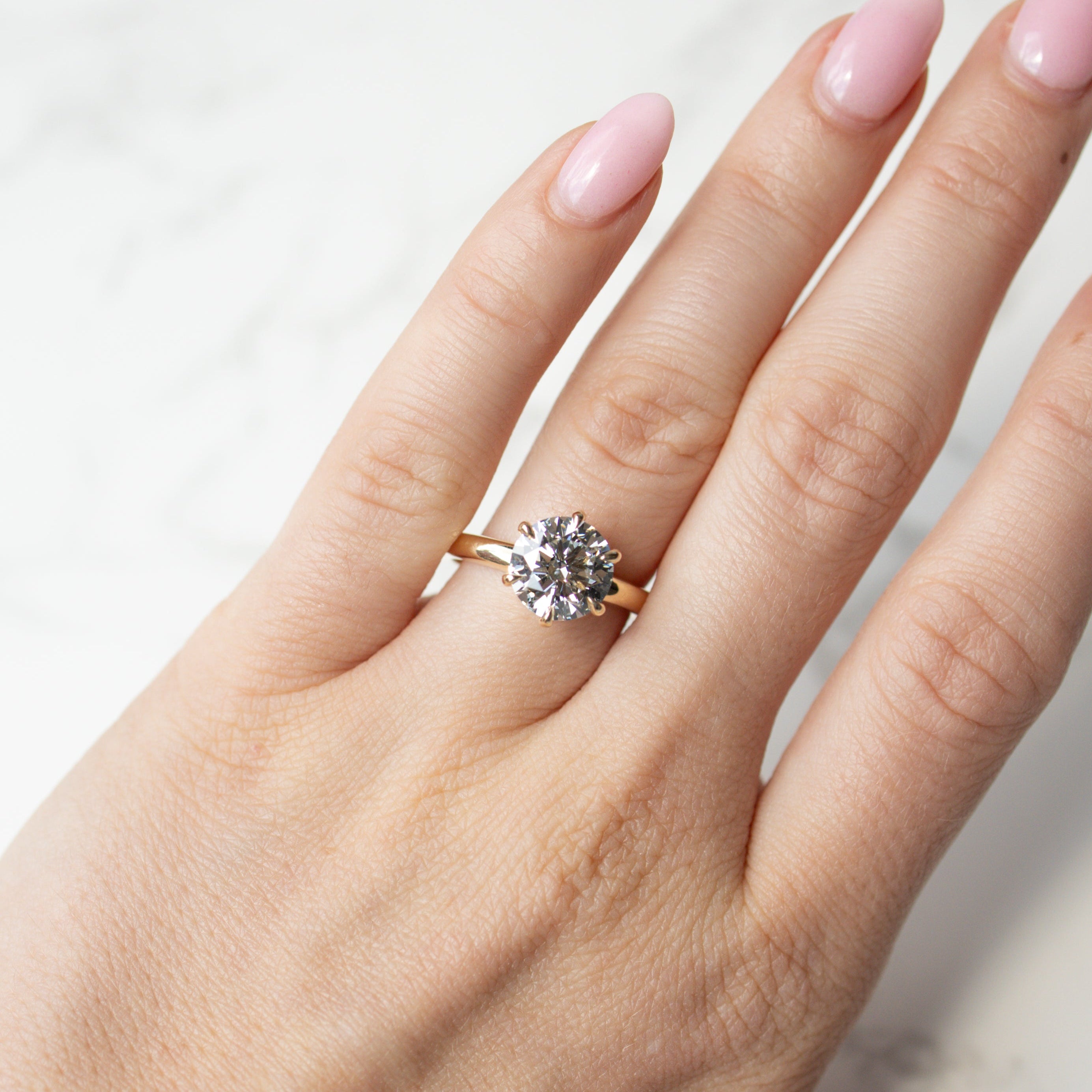A close-up of a hand with light pink manicured nails wearing a yellow gold ring with a clear round-cut diamond, set against a light marble background.