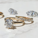 A yellow gold engagement ring and wedding band stacked on a white marble backdrop with a marquise and elongated cushion diamond in the background.