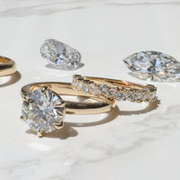 A yellow gold engagement ring and wedding band stacked on a white marble backdrop with a marquise and elongated cushion diamond in the background.
