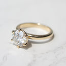A classic solitaire engagement ring featuring a round brilliant-cut diamond set in a yellow gold six-prong setting, resting on a white marble surface.