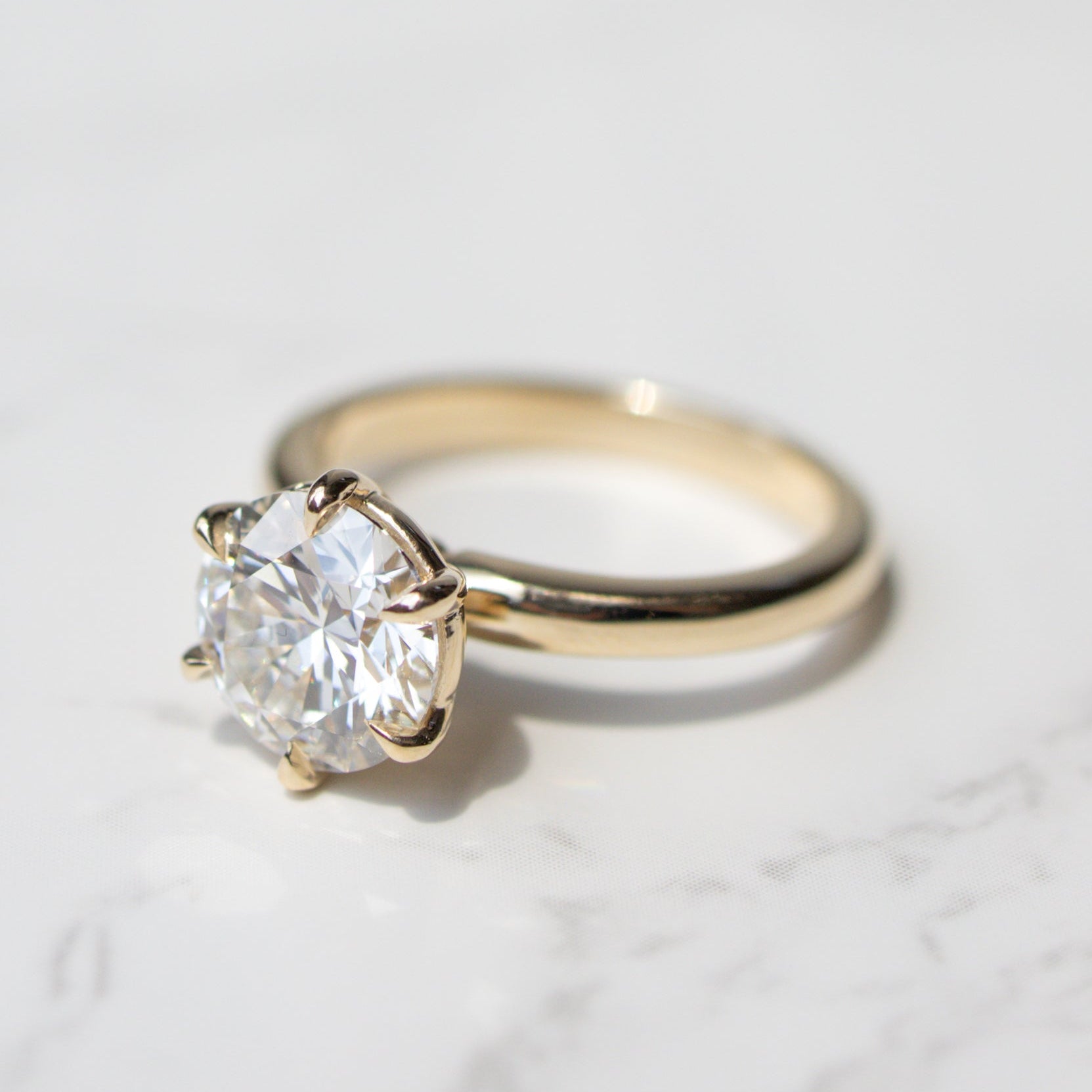 A classic solitaire engagement ring featuring a round brilliant-cut diamond set in a yellow gold six-prong setting, resting on a white marble surface.