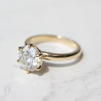 A classic solitaire engagement ring featuring a round brilliant-cut diamond set in a yellow gold six-prong setting, resting on a white marble surface.