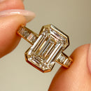 A close-up of a yellow gold engagement ring with a large emerald-cut diamond center stone and baguette side stones, held between fingers against a soft beige background.