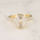 Macro shot of a yellow gold half bezel engagement ring with an emerald cut diamond center on a white marble backdrop