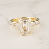 Macro shot of a yellow gold half bezel engagement ring with an emerald cut diamond center on a white marble backdrop