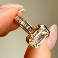 A close-up of a gold ring with a large emerald-cut diamond center stone and baguette side stones, held between fingers against a soft beige background.