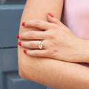 A close-up lifestyle shot of a person's hand resting on their arm, showcasing a gold ring with a moval-cut diamond. The ring features a yellow gold band with a georgian setting.