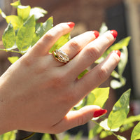 A gold engagement ring with an east west set moval diamond worn on a person's ring finger, shown in a lifestyle setting with green leaves and red nail polish in the background.