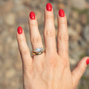 A close-up of a hand with bright red nail polish wearing a gold ring with a moval-cut diamond, set against a blurred outdoor background of rocks and soil.