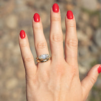 A close-up of a hand with bright red nail polish wearing a gold ring with a moval-cut diamond, set against a blurred outdoor background of rocks and soil.