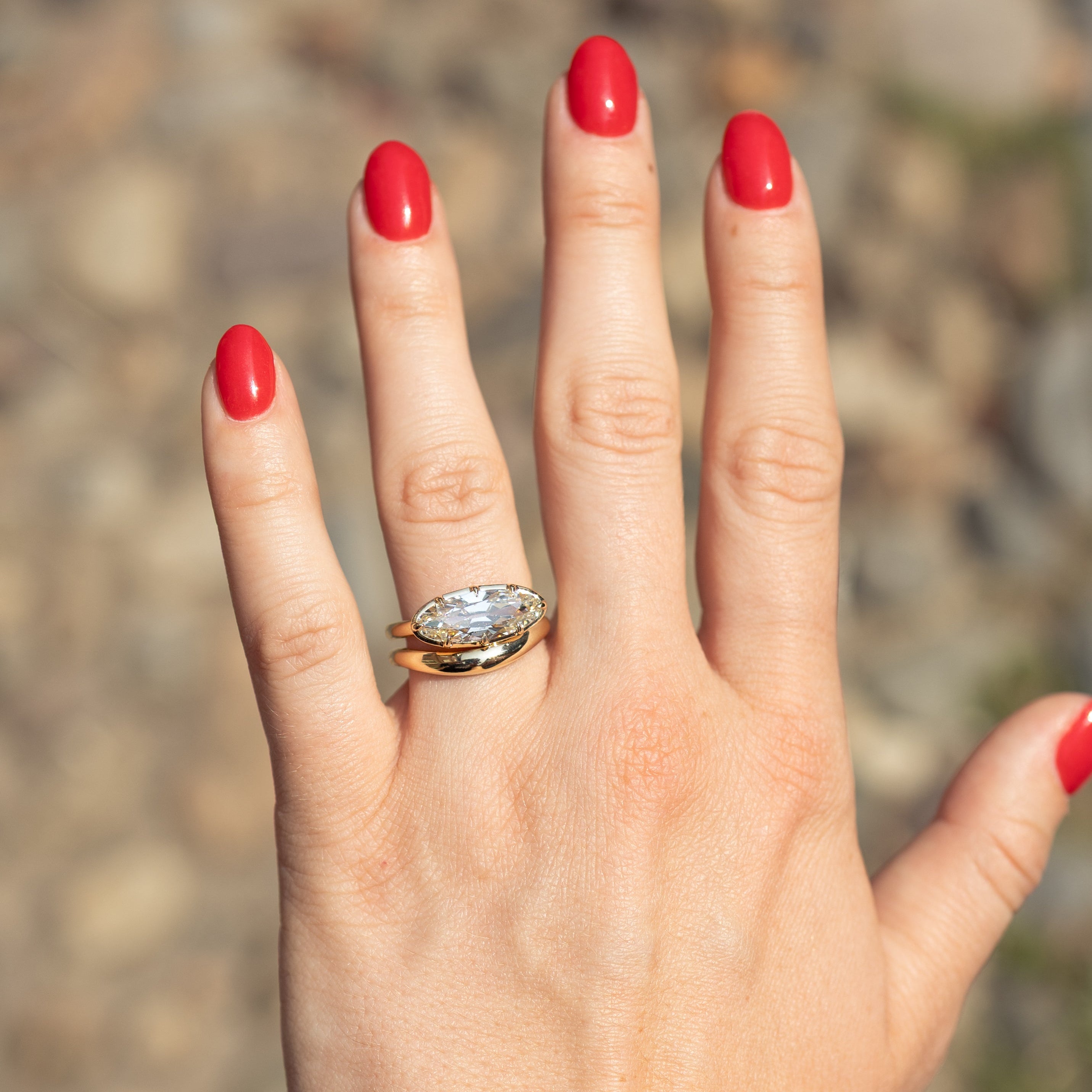 A close-up of a hand with bright red nail polish wearing a gold ring with a moval-cut diamond, set against a blurred outdoor background of rocks and soil.