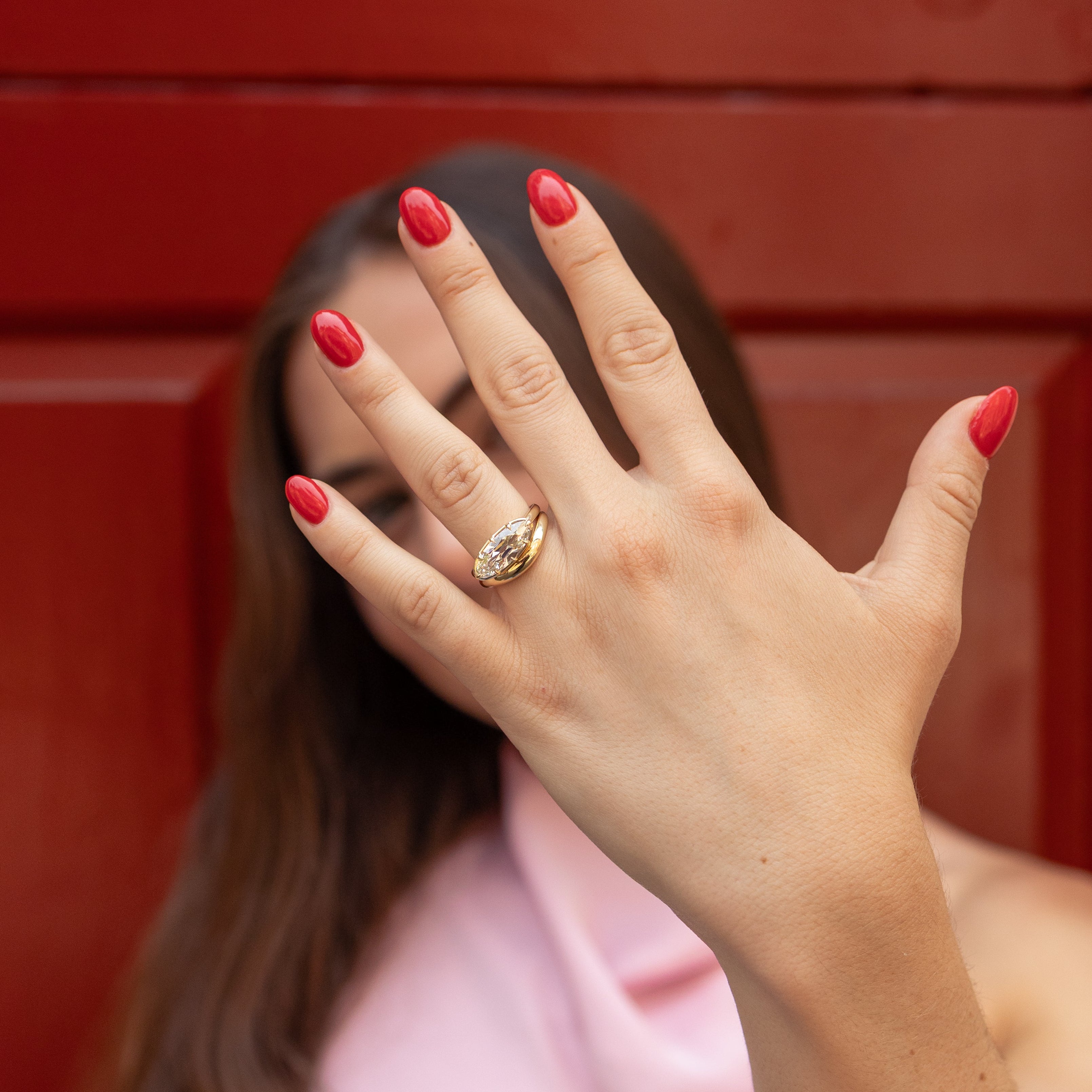 A woman with long dark hair and red nail polish holds her hand up in front of her face, wearing a gold engagement ring with an east west moval diamond on her ring finger. She is wearing a light pink top against a dark red wooden door background.