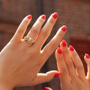A close-up of two hands with bright red nail polish, one wearing a gold moval diamond engagement ring, set against a blurred brick building background.
