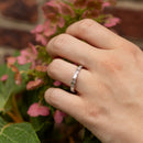 A close-up of a hand wearing a silver band ring with rectangular baguette-cut stones, set against a soft-focus background of pink hydrangea flowers and green leaves.