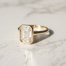 A gold bezel style engagement ring featuring an emerald-cut diamond, resting on a white marble surface with soft shadows.
