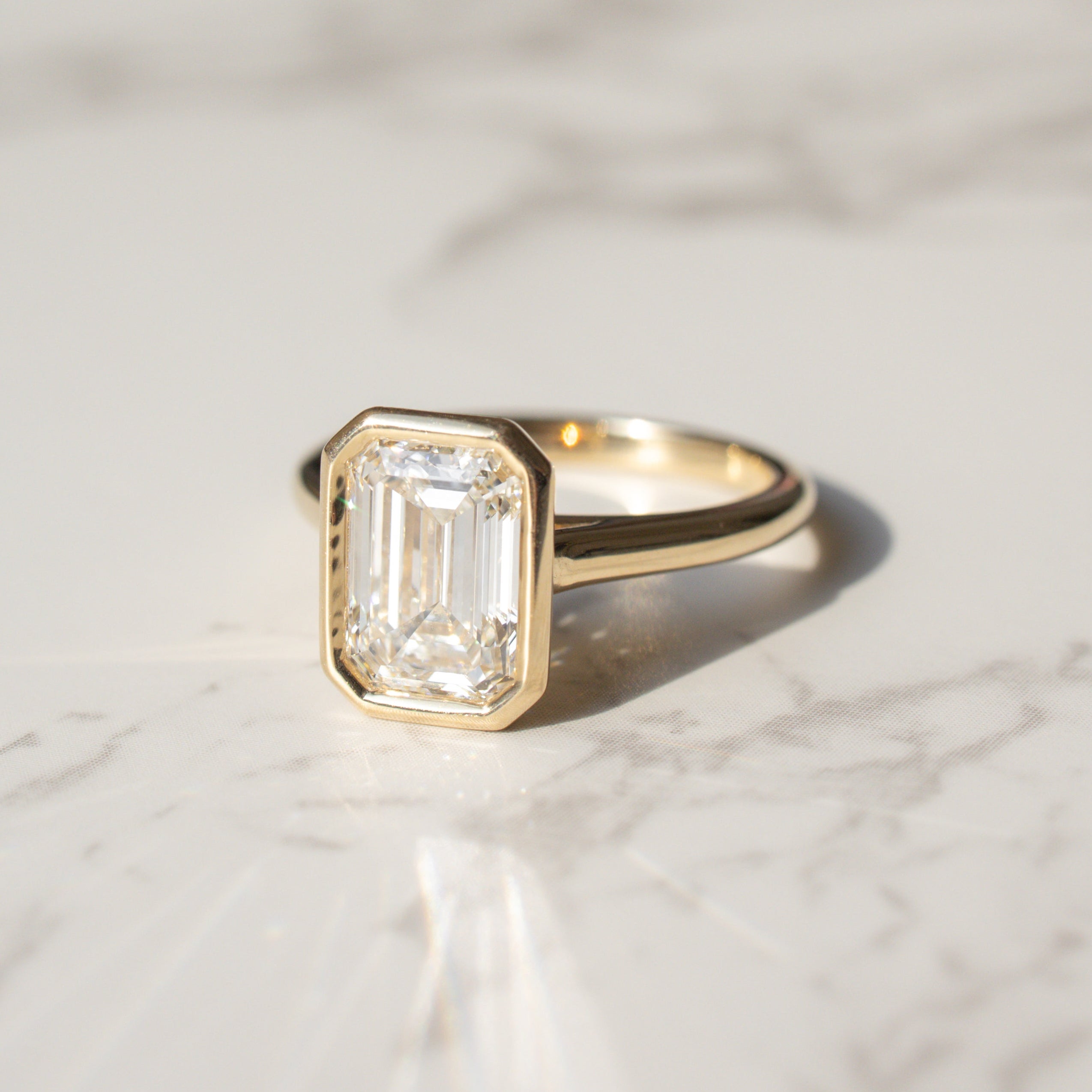 A gold bezel style engagement ring featuring an emerald-cut diamond, resting on a white marble surface with soft shadows.