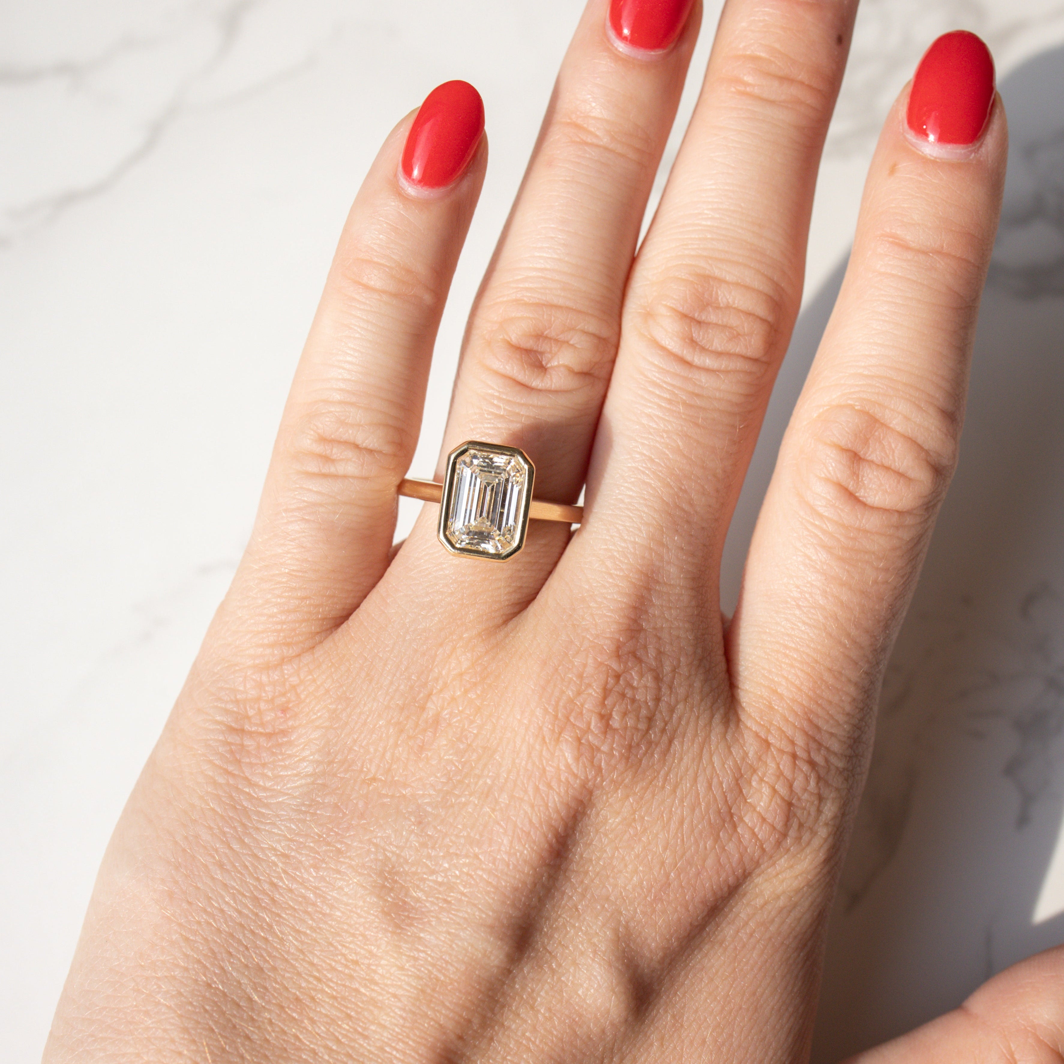 A close-up of a hand with bright red nail polish wearing a gold bezel ring with an emerald-cut diamond, set against a white marble background.