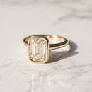 A gold bezel engagement ring with an emerald-cut clear diamond, resting on a white marble surface with soft shadows.