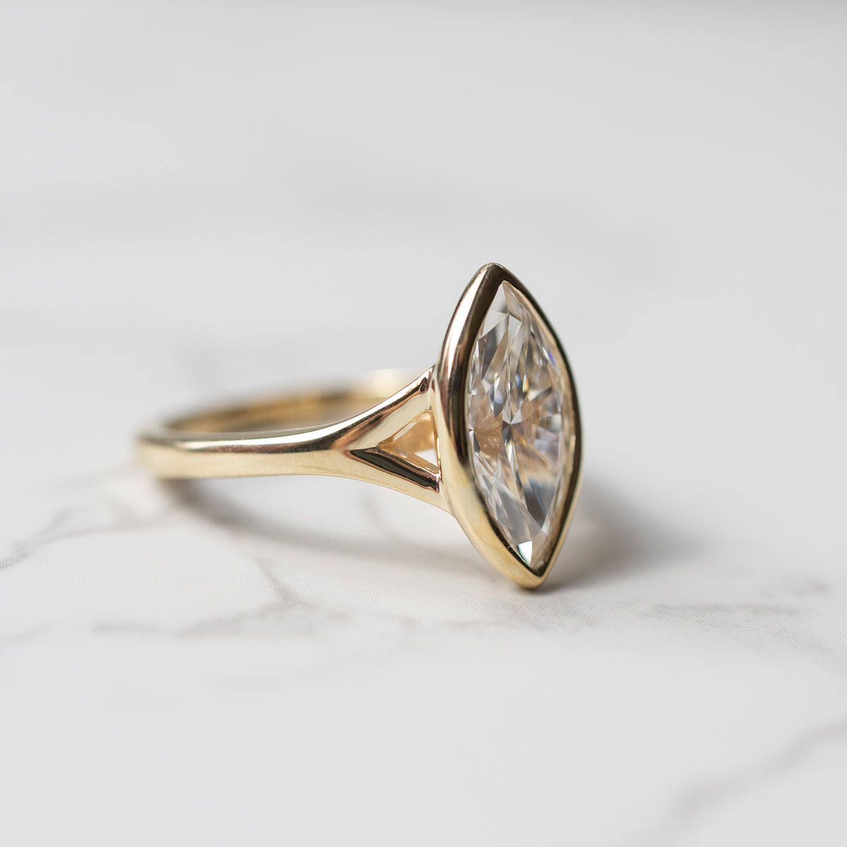 Side angle view of the split shank on an engagement ring setting. The ring is yellow gold and the diamond is a marquise. It's sitting on a white marble backdrop.