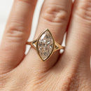 A large marquise cut diamond engagement ring set in yellow gold on the left ring finger of a woman's hand. In the background is a blurry white backdrop.