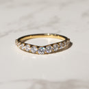 A gold band ring with a row of clear pavé-set stones, resting on a white marble surface with a blurred dark background.
