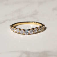 A gold band ring with a row of clear pavé-set stones, resting on a white marble surface with a blurred dark background.