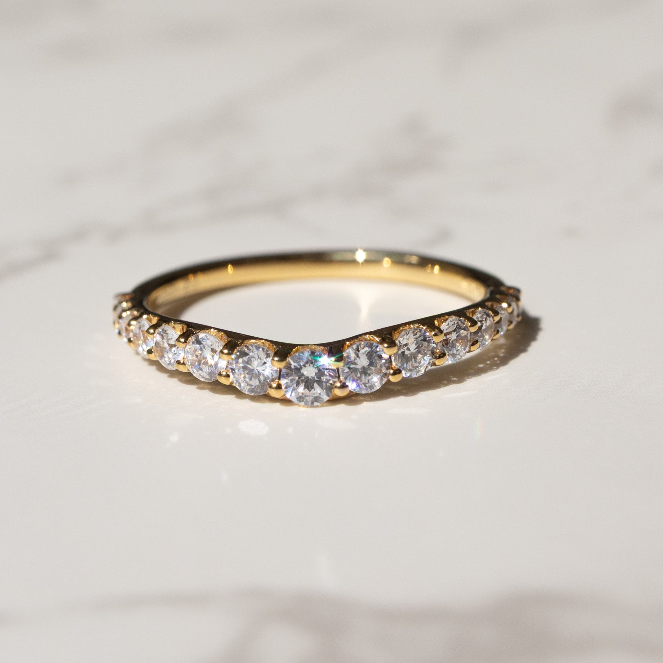 A gold band ring with a row of clear pavé-set stones, resting on a white marble surface with a blurred dark background.