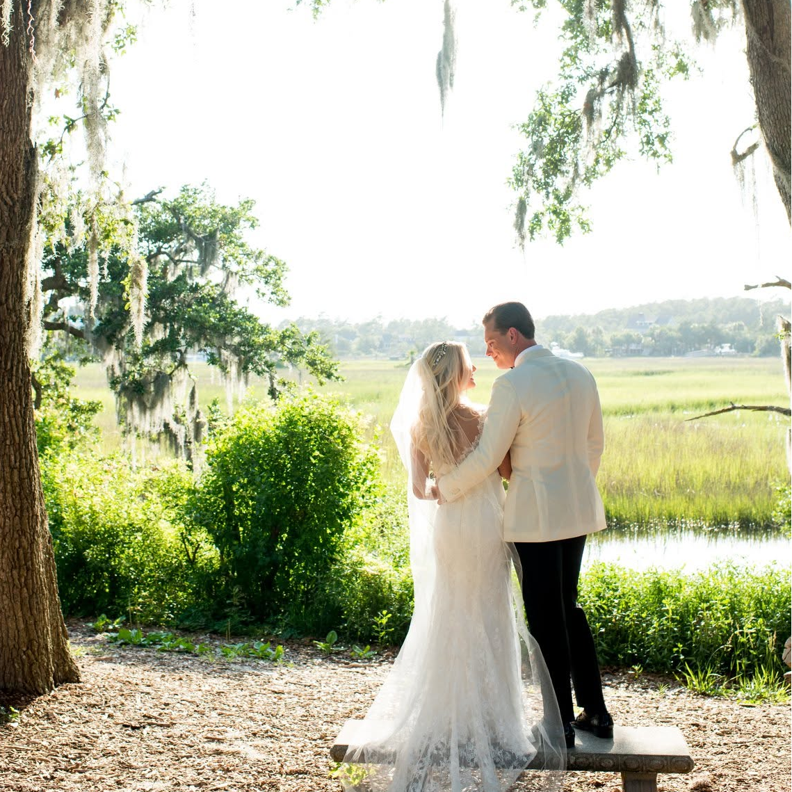 A bride in a white lace wedding gown and veil and a groom in a light suit stand together on a bench beneath a large tree draped with Spanish moss, with a sunlit marsh landscape in the background.