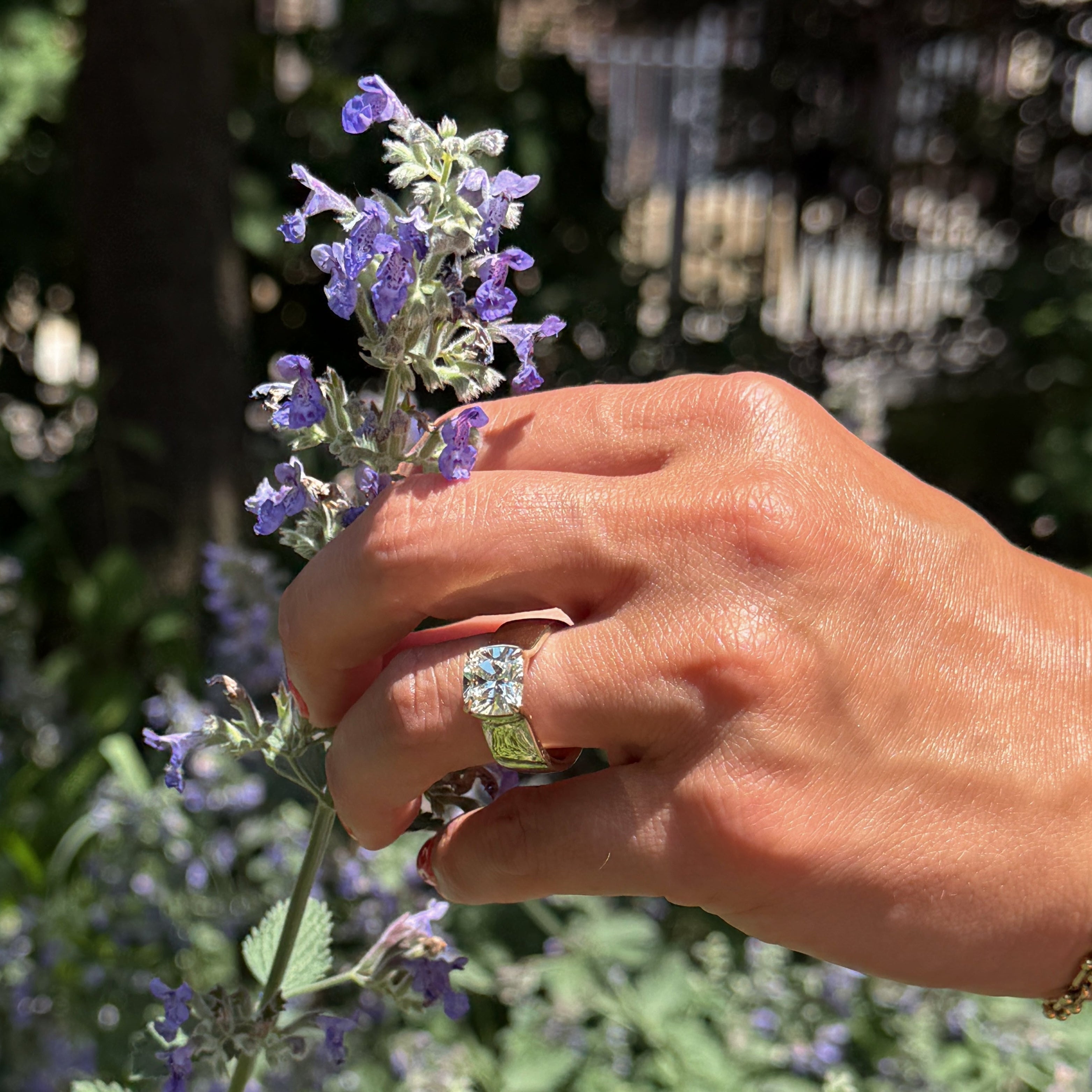 A large cushion cut engagement ring on a left ring finger. The hand is holding purple flowers.