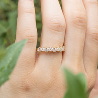 A hand being held up against green foliage. On the left ring finger of the hand is a gold diamond band with marquise and round diamonds clustered together.