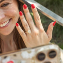 A close-up lifestyle shot of a woman showing off a gold diamond ring on her finger, with her hand raised in front of a phone camera. She has bright red nail polish, and the background is a soft-focus outdoor setting.