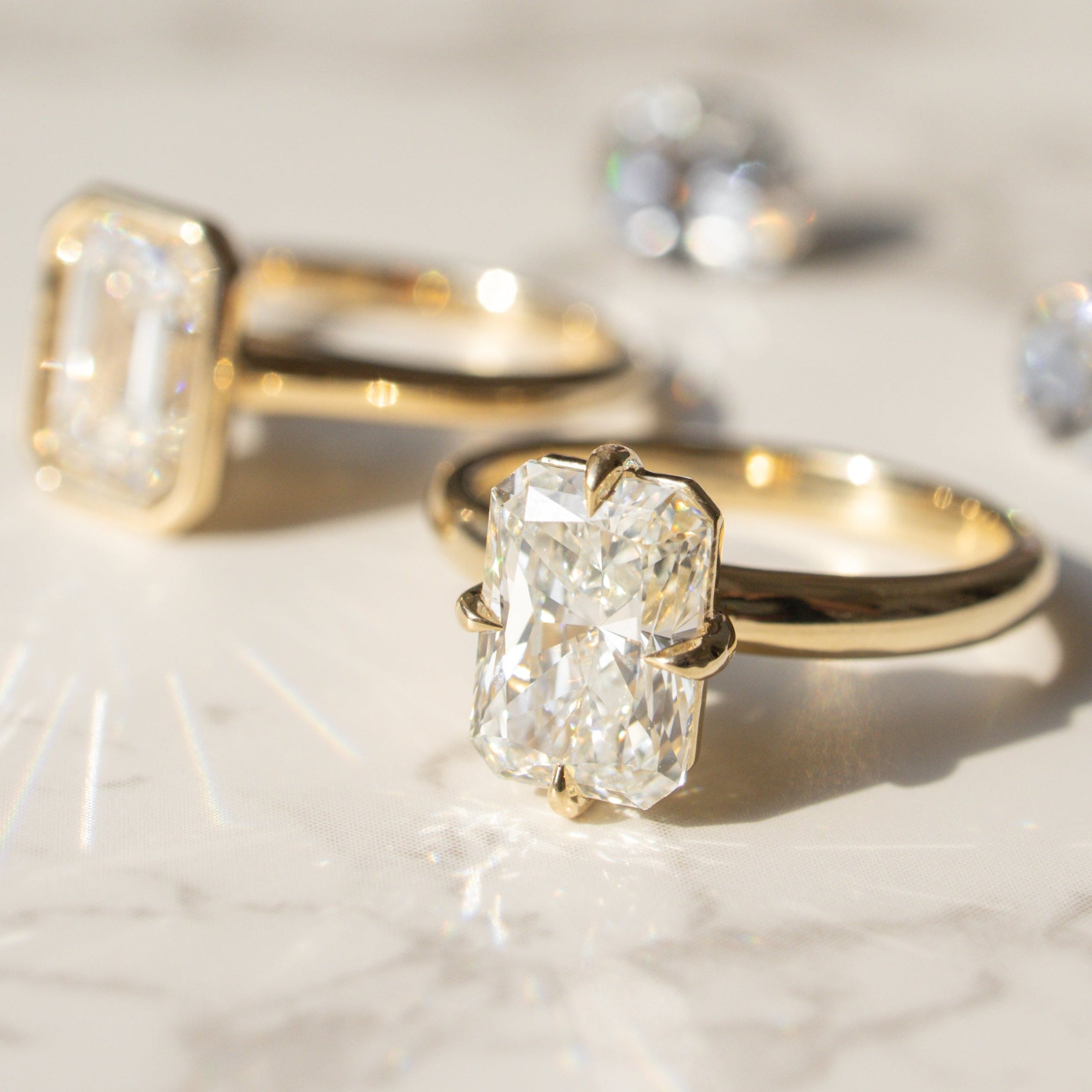 Two yellow gold rings with clear diamonds are arranged on a light marble surface. The front ring features a large rectangular radiant-cut diamond, while the ring behind it has an emerald-cut diamond set in a bezel.