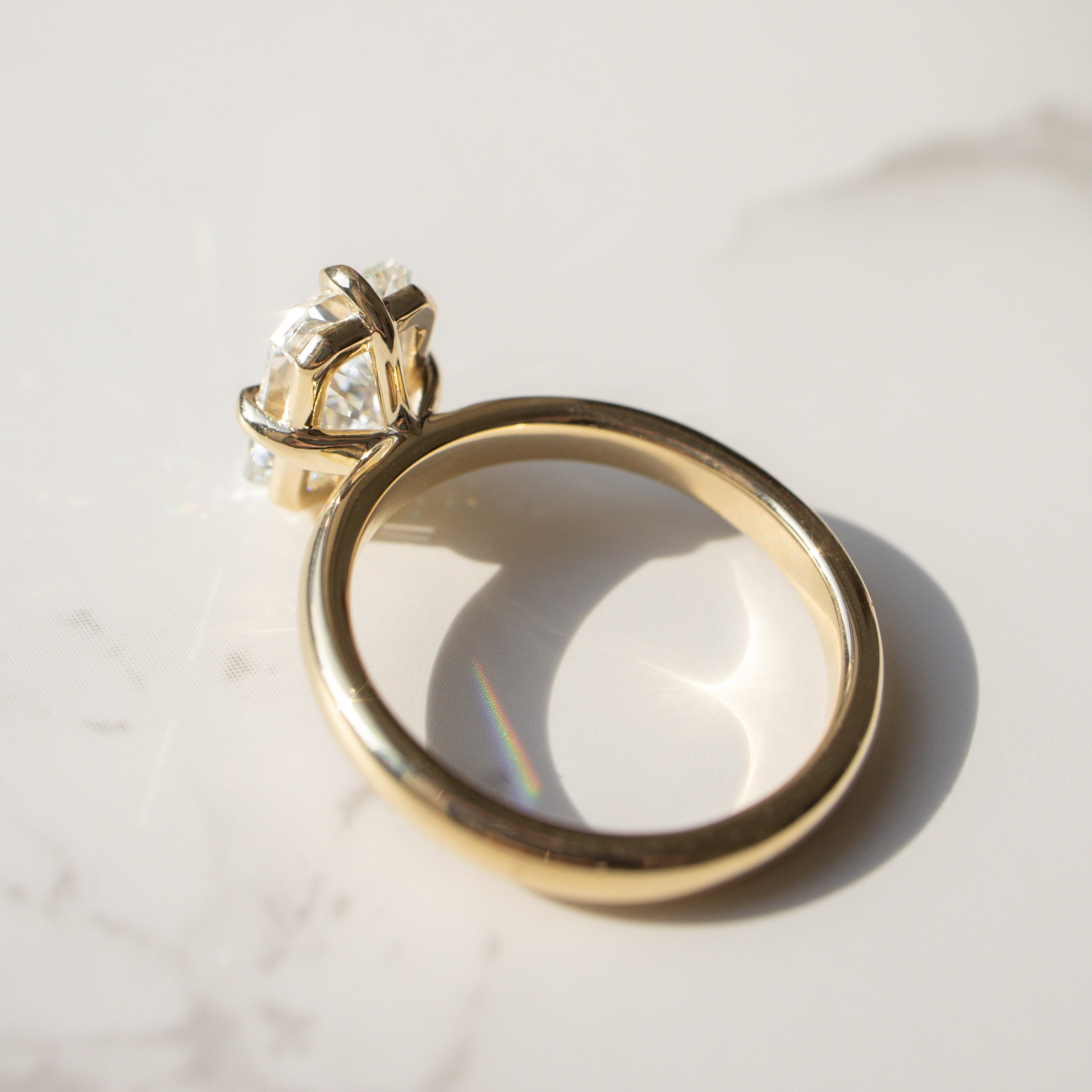 A gold ring with a radiant diamond in a compass prong setting, resting on a white marble surface with a soft shadow.