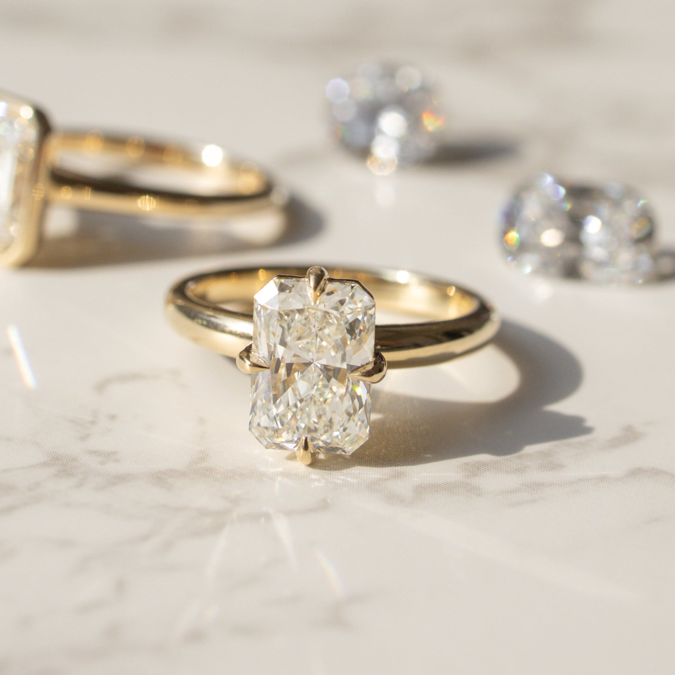 Two gold engagement rings with loose diamonds behind them arranged on a light marble surface in bright sunlight.