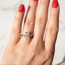A close-up of a hand with bright red nail polish wearing a yellow gold ring with a radiant cut diamond, set against a light marble background.