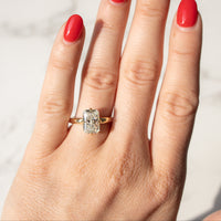 A close-up of a hand with bright red nail polish wearing a yellow gold ring with a radiant cut diamond, set against a light marble background.
