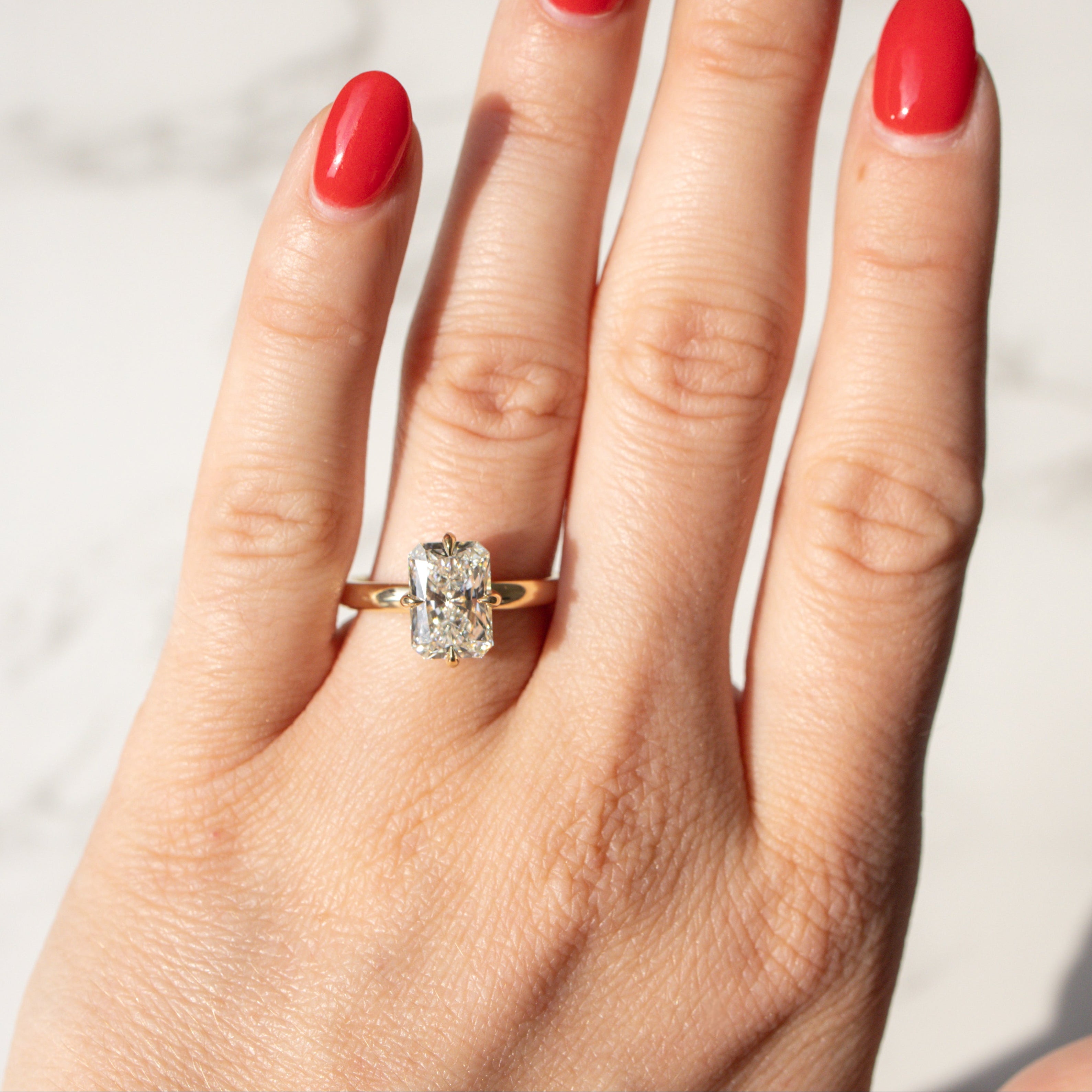A close-up of a hand with bright red nail polish wearing a yellow gold ring with a radiant cut diamond, set against a light marble background.