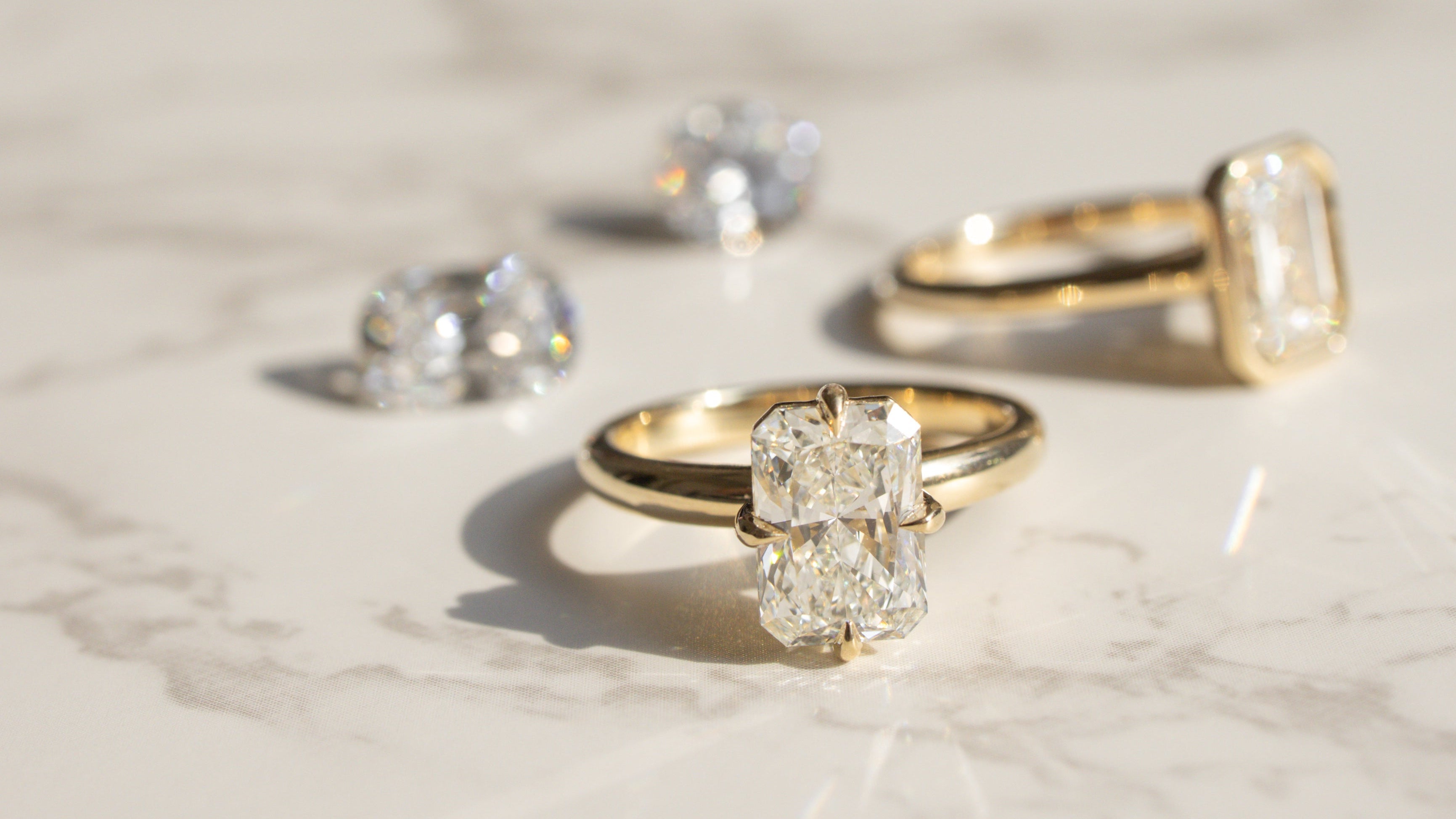 Two gold engagement rings and loose diamonds arranged on a light marble surface in bright sunlight.