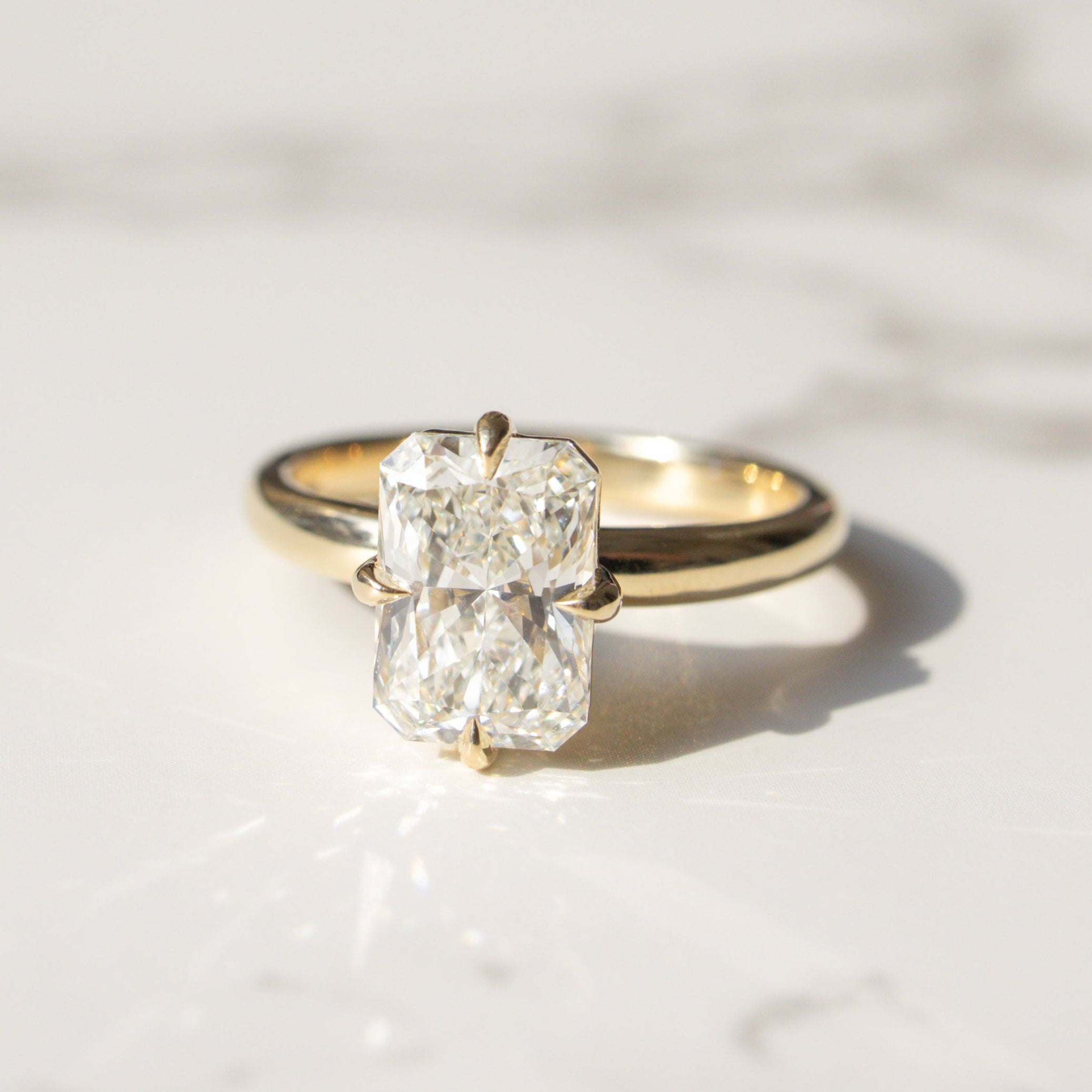 A yellow gold ring with a large rectangular radiant-cut diamond solitaire, resting on a white marble surface with soft shadows.