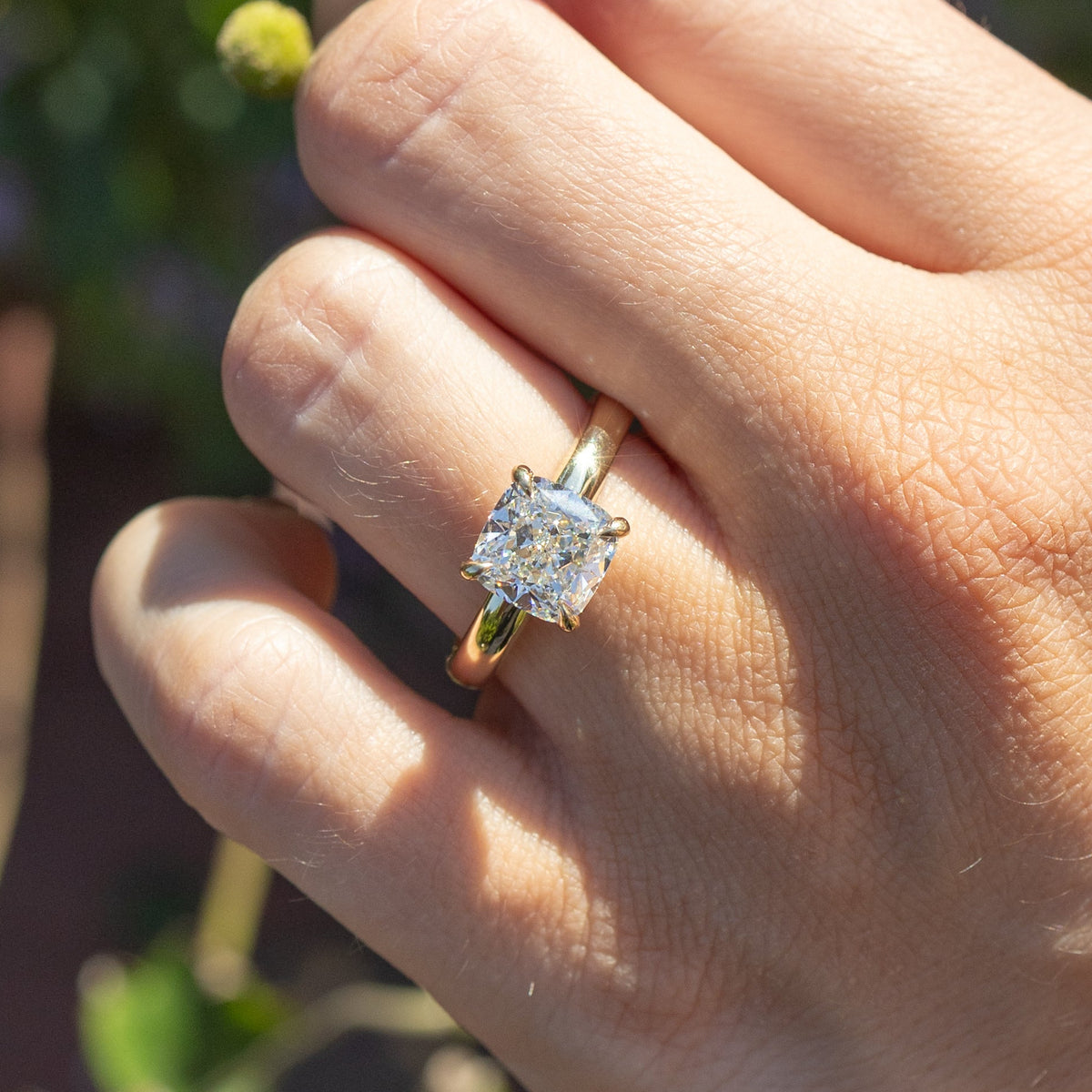 A close up shot of a hand holding a pink flower. On the hand's left ring finger is a large yellow gold engagement ring with a large cushion cut diamond in the center.