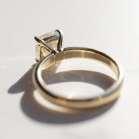 The back profile of a yellow gold engagement ring sitting on a white marble backdrop. The ring has a large cushion cut diamond in the middle.
