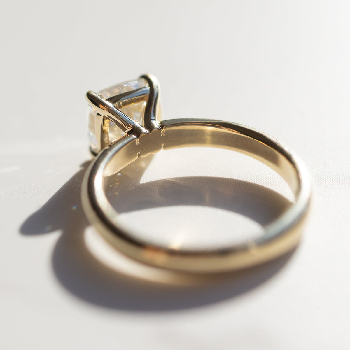 The back profile of a yellow gold engagement ring sitting on a white marble backdrop. The ring has a large cushion cut diamond in the middle.