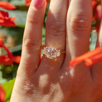 A sparkling engagement ring on a hand in the middle of red flowers. The ring has a cushion cut center diamond with cadillac side stones.