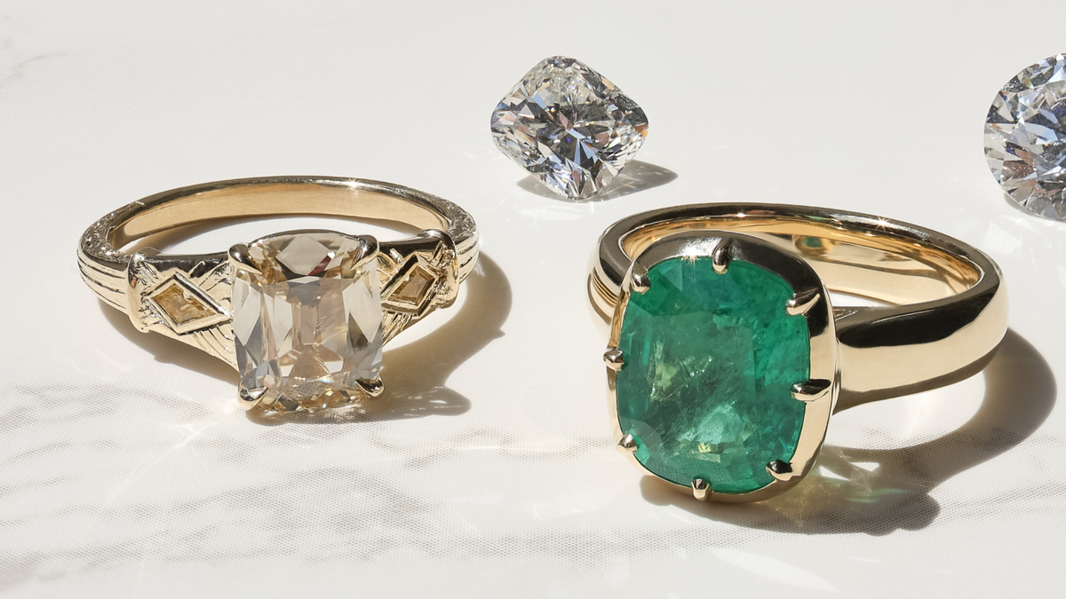 Two yellow gold custom engagement rings sitting on a white marble backdrop in sunlight. One ring has a warm natural diamond, the other has a green emerald gemstone. There are two loose diamonds behind them.