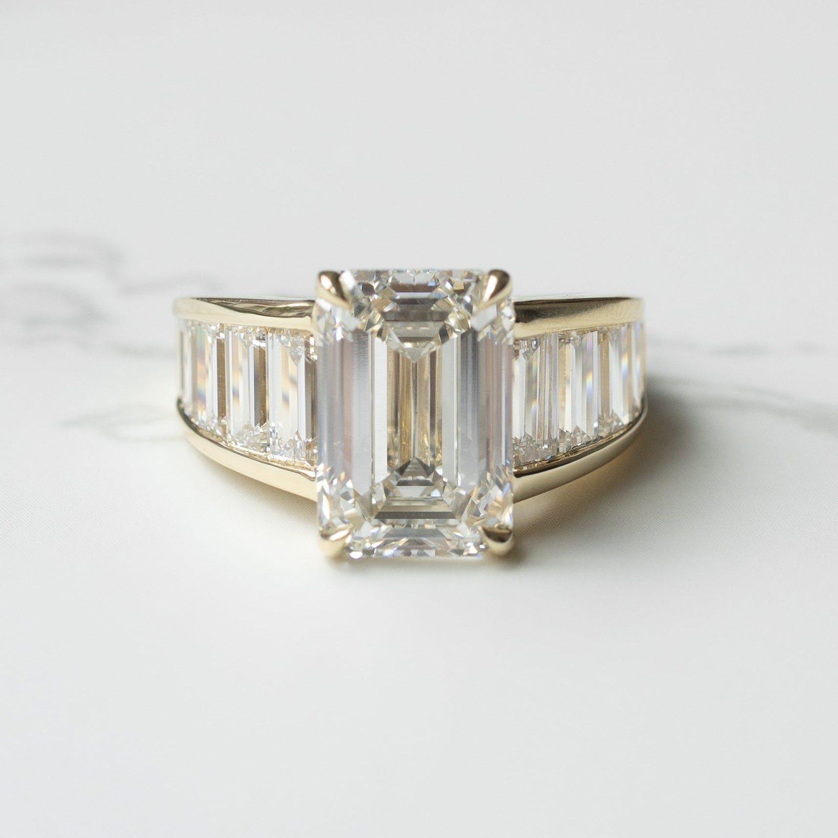 A large emerald cut diamond set in a yellow gold setting on a white marble backdrop with shadows. The ring has baguette diamonds tapering in size down each side of the band.