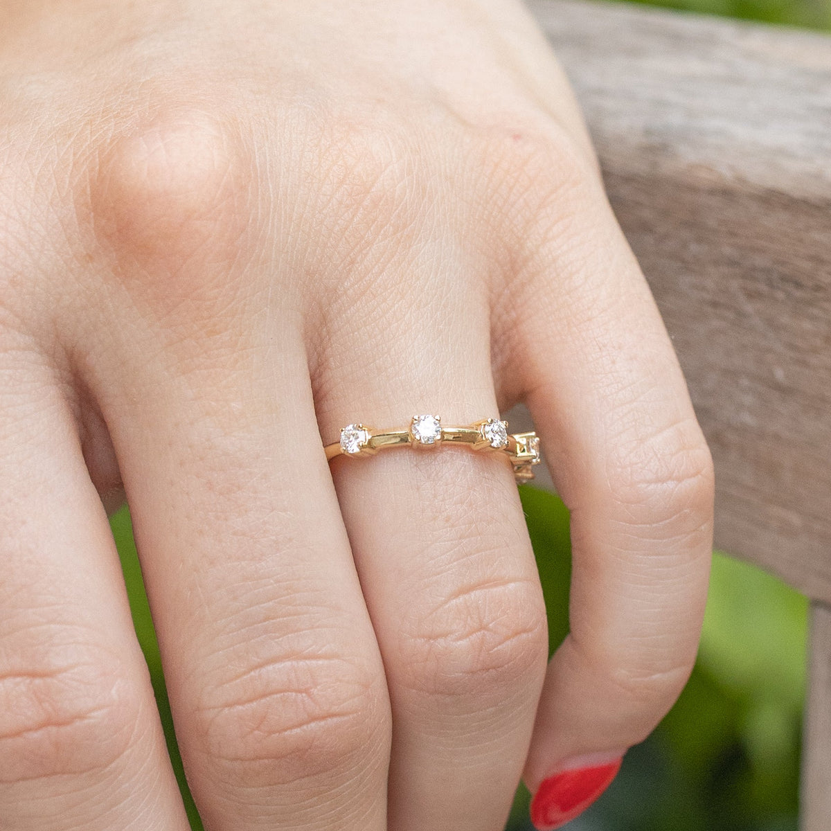 A hand against the side of a wooden bench. On the left ring finger of the hand is a yellow gold wedding band with stationed round diamonds.