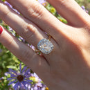 A close up shot of a large elongated cushion diamond engagement ring. The center diamond is surrounded by a halo of small prong-set round diamonds. In the background is a hand and blurry purple and green flowers.